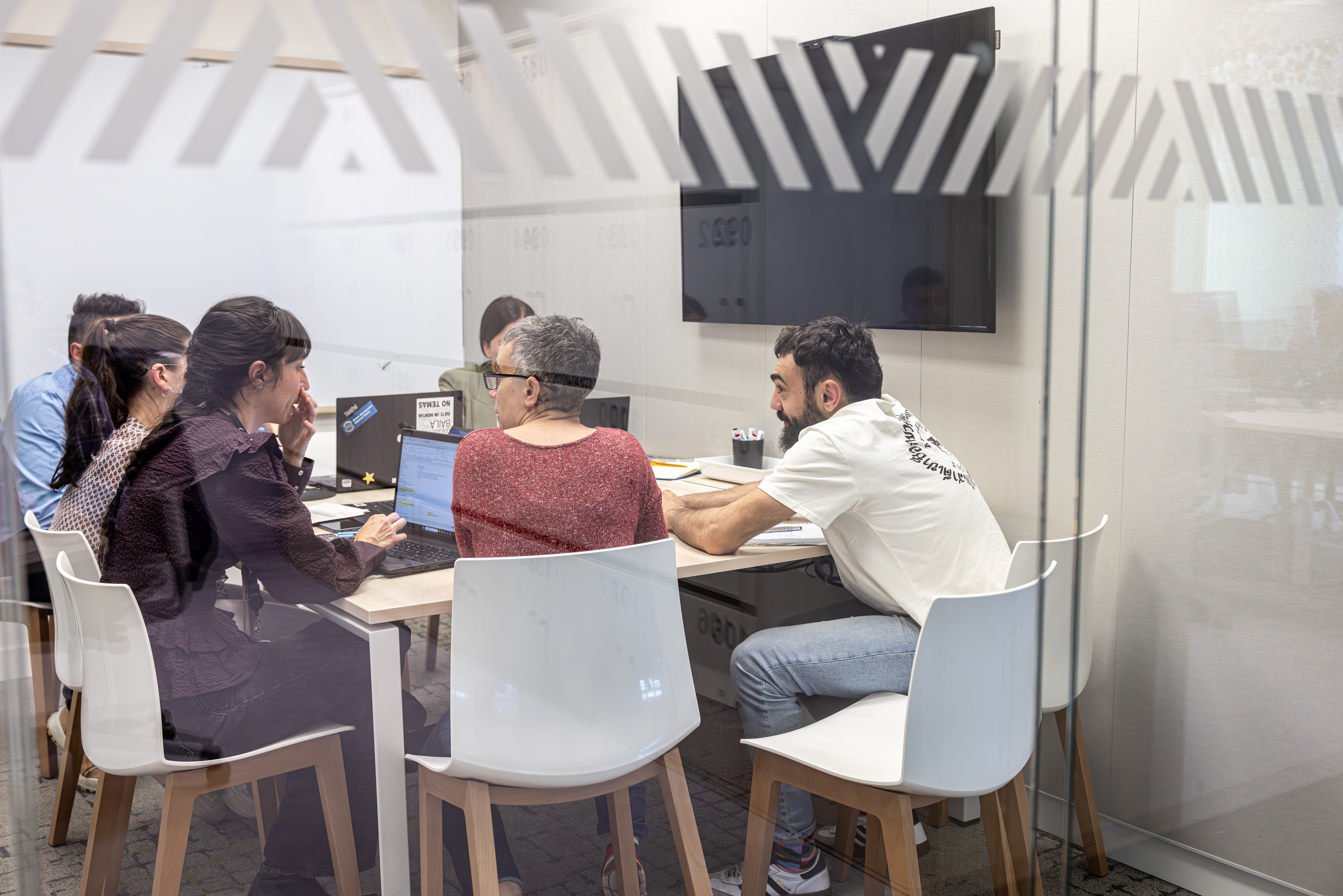 Equipo reunido en una sala de reuniones con paredes de cristal, trabajando con portátiles y documentos alrededor de una mesa