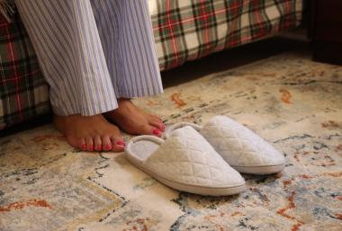 Fotografía de la campaña #NadieSinZapatillas, en la que se ven los pies de una persona entre una cama y las pantuflas solidarias de Hogar Sí