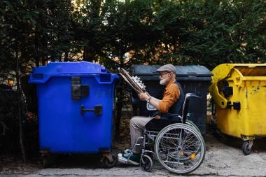 Un hombre mayor en silla de ruedas se dispone a tirar restos no orgánicos en un contenedor específico