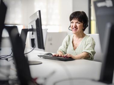 Fotografía de una mujer con auriculares y micrófono trabaja frente a un ordenador en una oficina luminosa, con las manos sobre el teclado y varios monitores alrededor.