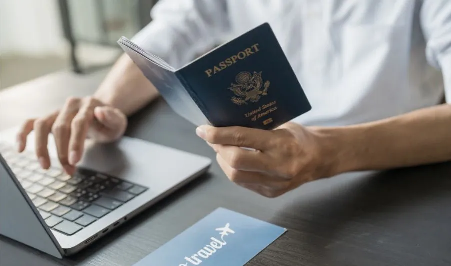 Hombre aguantando un pasaporte y planificando un viaje con su portátil