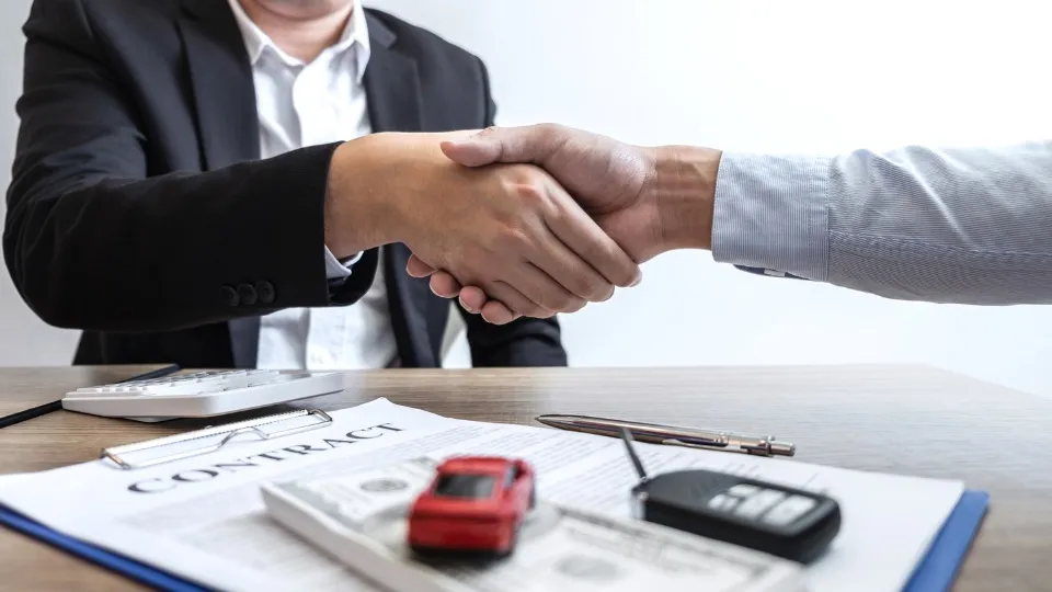 Dos personas se dan la mano sobre una mesa de madera con un contrato, una calculadora, un bolígrafo, un coche de juguete y las llaves del coche.