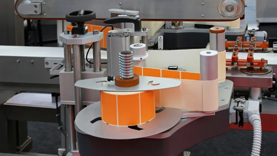 Primer plano de una máquina de impresión de etiquetas con un rollo de etiquetas naranja, componentes metálicos y una cinta transportadora, que transmite eficacia y precisión industrial.