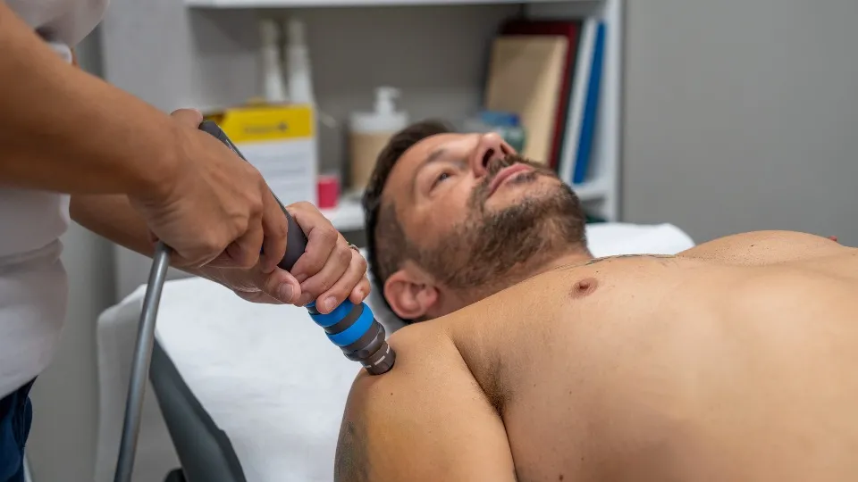 Las manos de un fisioterapeuta aplicando la técnica de ondas de choque en el hombro de un hombre que está tumbado en una camilla. 