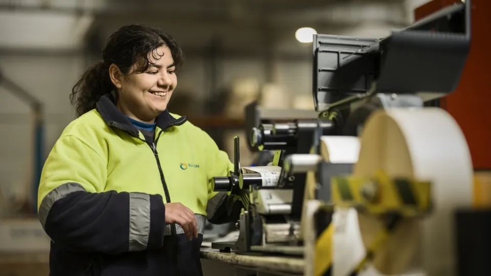 Una mujer, empleada de ILUNION, con chaqueta de alta visibilidad sonríe mientras maneja maquinaria de impresión de etiquetas en un entorno industrial. 