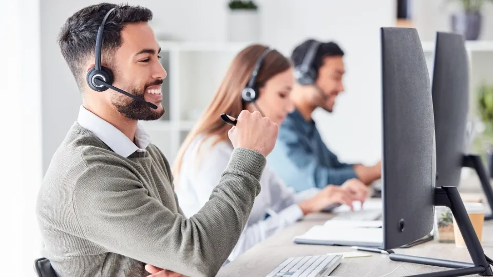 Hombre sonriendo y usando auriculares, sentado en un escritorio con un ordenador. Al fondo, dos compañeros están concentrados, creando un ambiente de oficina positivo.