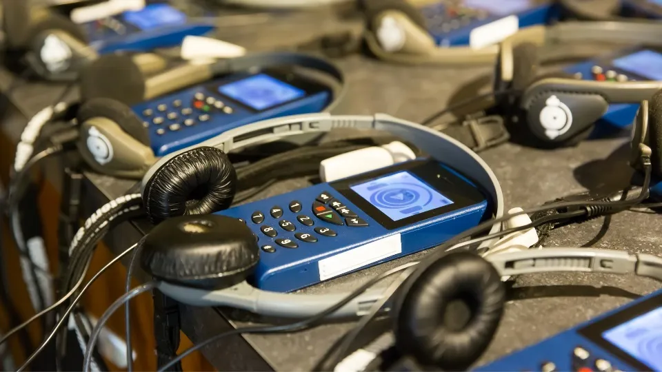 Primer plano de un grupo de audioguías azules con auriculares sobre una mesa, listas para usar. Las pantallas de los dispositivos muestran un icono de reproducción, creando un ambiente organizado y funcional.