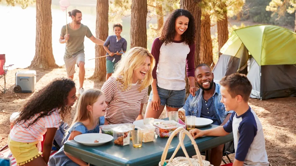 Un grupo de amigos y niños disfrutan de un picnic en un campamento forestal, sonriendo y riendo. Una tienda de campaña y árboles al fondo transmiten alegría.