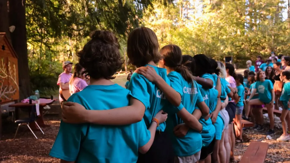Un grupo de campistas con camisetas verde azulado se encuentran cogidos del brazo en un pintoresco entorno forestal. El ambiente es animado y alegre, con otros reunidos cerca.