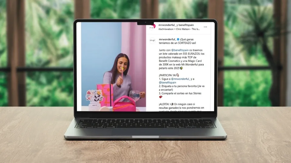 Una laptop sobre una mesa de madera muestra una publicación de Instagram. Una mujer sonríe mientras sostiene una brocha de maquillaje, con cajas y bolsas de colores cerca. Refleja una colaboración de Mr Wonderful con una marca de cosmética llamada Benefit.