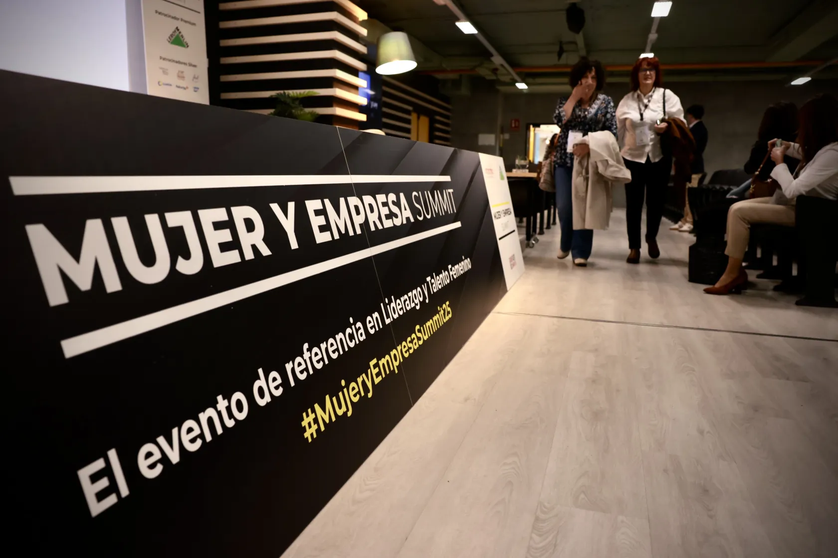 Dos mujeres conversan al fondo. De izquierda a derecha, un panel indica que están en el salón Mujer y Talento, organizado por Intrama