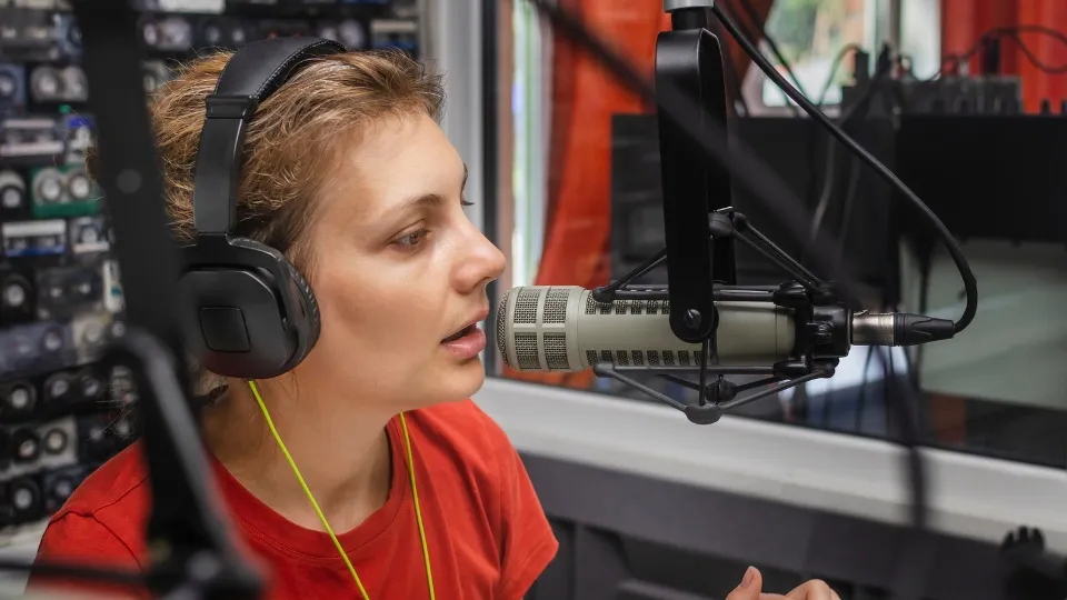 Una persona con auriculares habla por un micrófono en un estudio. Lleva una camisa roja y parece concentrada. Al fondo hay equipos y cables.