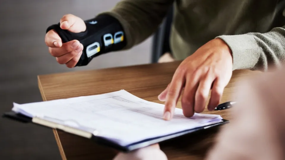 Una persona con una muñequera revisa un documento en un portapapeles sobre una mesa de madera. Otra mano señala el papel, indicando una conversación concentrada.