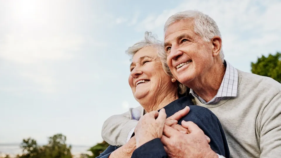 Una pareja de ancianos se abraza alegremente bajo un cielo radiante, irradiando calidez y satisfacción. Los árboles y la luz del sol crean un ambiente sereno y alentador.