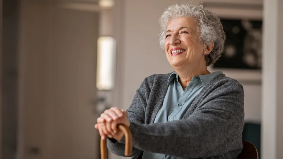 Una mujer mayor con una cálida sonrisa, está sentada en el interior con las manos apoyadas en un bastón. Lleva un cárdigan gris, creando una atmósfera de paz.