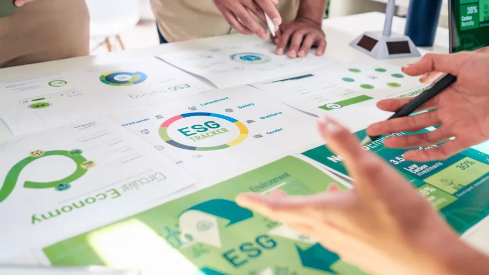 Varias manos señalan y discuten gráficos ambientales sobre una mesa, con el foco puesto en un rastreador ESG. La escena evoca trabajo en equipo y análisis.
