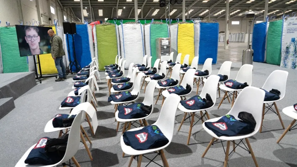 Filas de sillas blancas se disponen en un espacio industrial, cada una con una bolsa de tela de marca. Un hombre ajusta una pantalla que muestra a una persona. Al fondo, se ven barreras de colores.