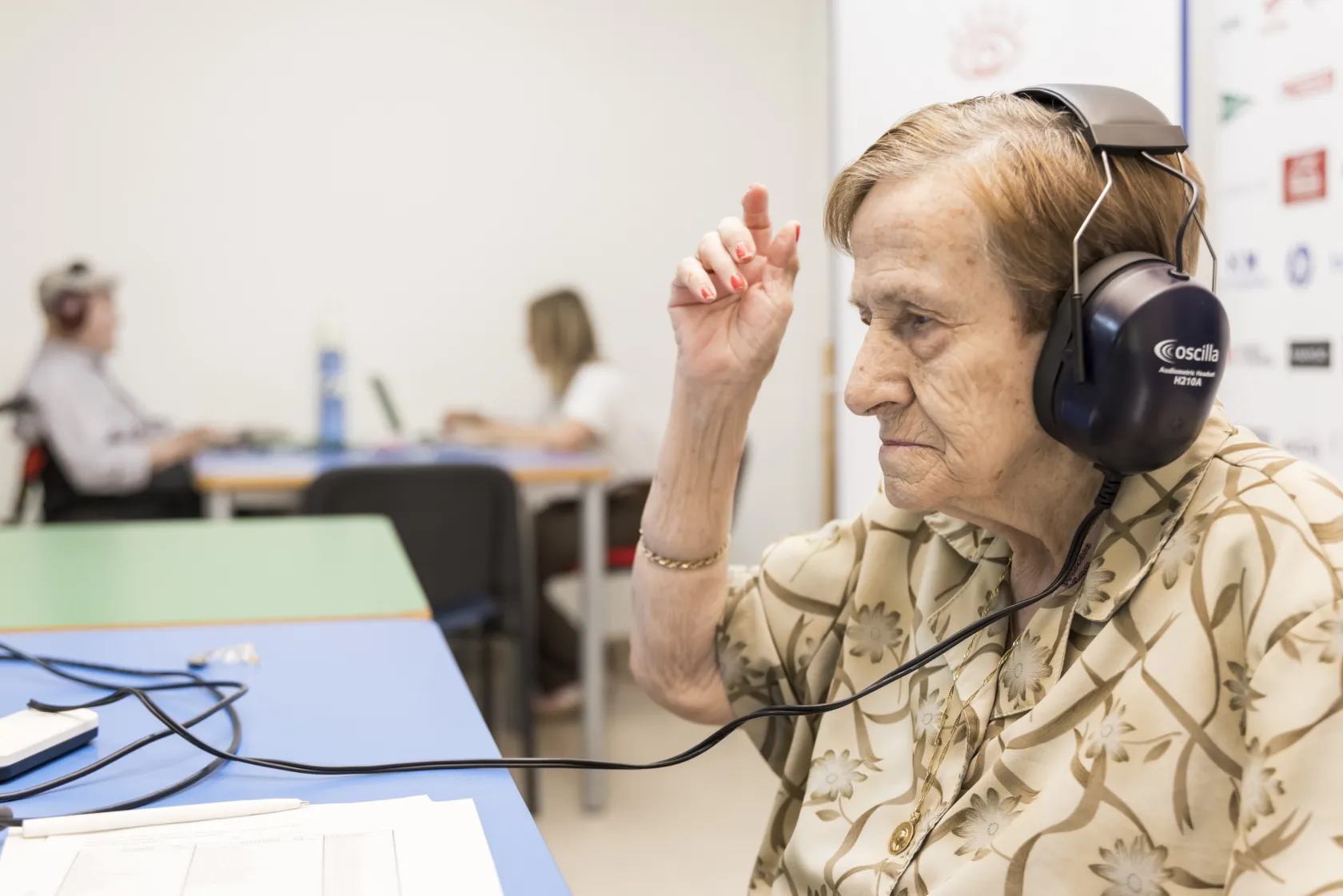 Una residente de ILUNION VidaSénior en Sonseca, que lleva unos grandes auriculares puestos, levanta la mano. Al fondo, otra persona mayor, un hombre, también está realizándose una prueba audiológica.