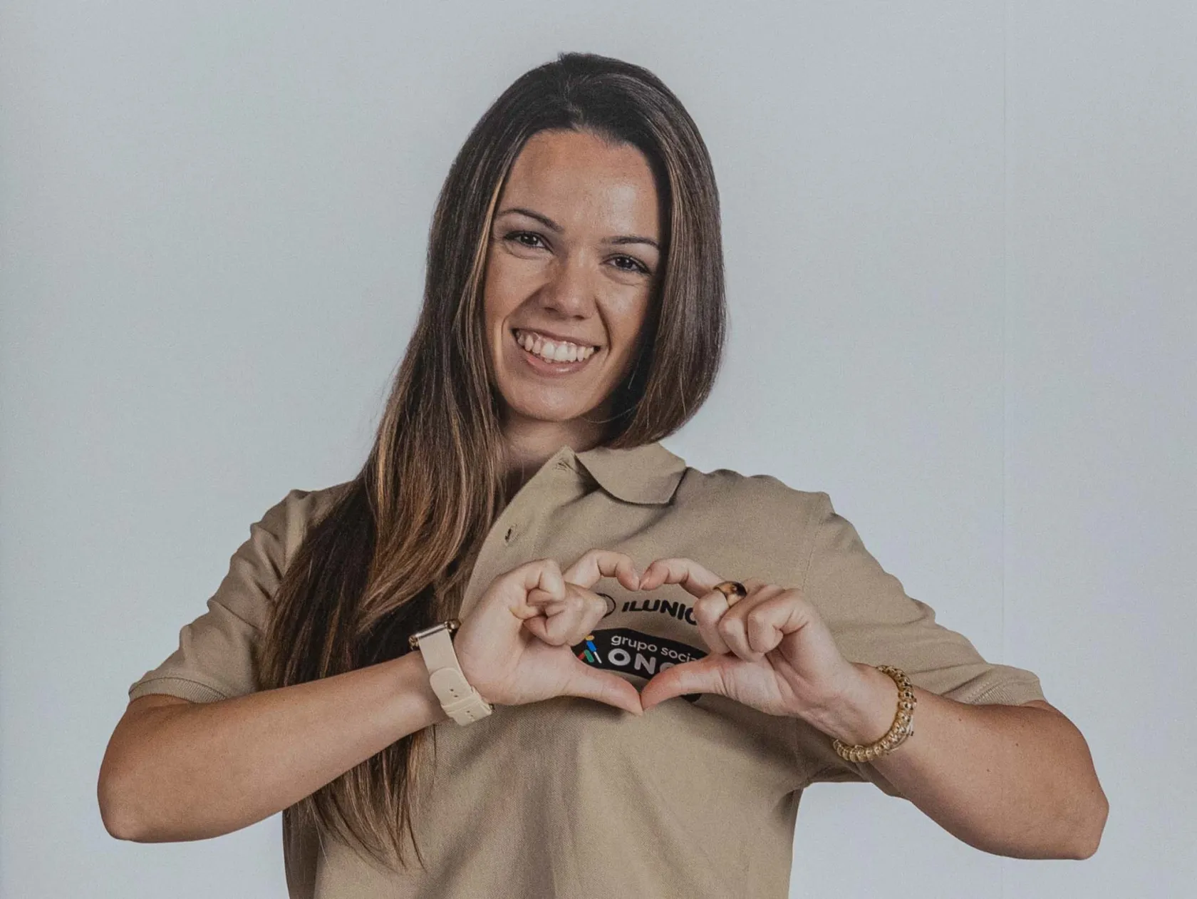 Una mujer joven y sonriente, vestida con el uniforme de ILUNION TuTienda, hace forma un corazón con sus manos