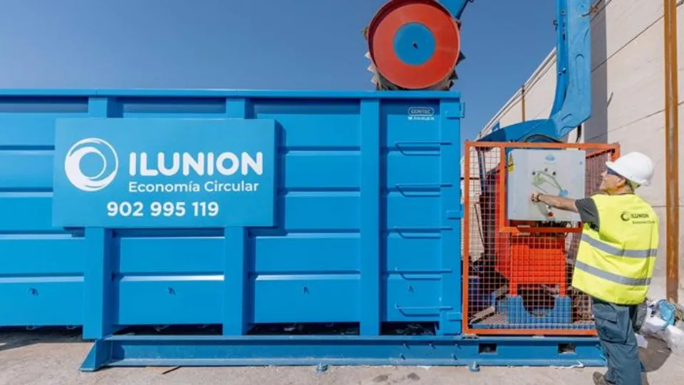 Un trabajador con casco y chaleco amarillo opera un panel de control en una gran máquina industrial azul marcada "ILUNION Economía Circular" bajo un cielo despejado.
