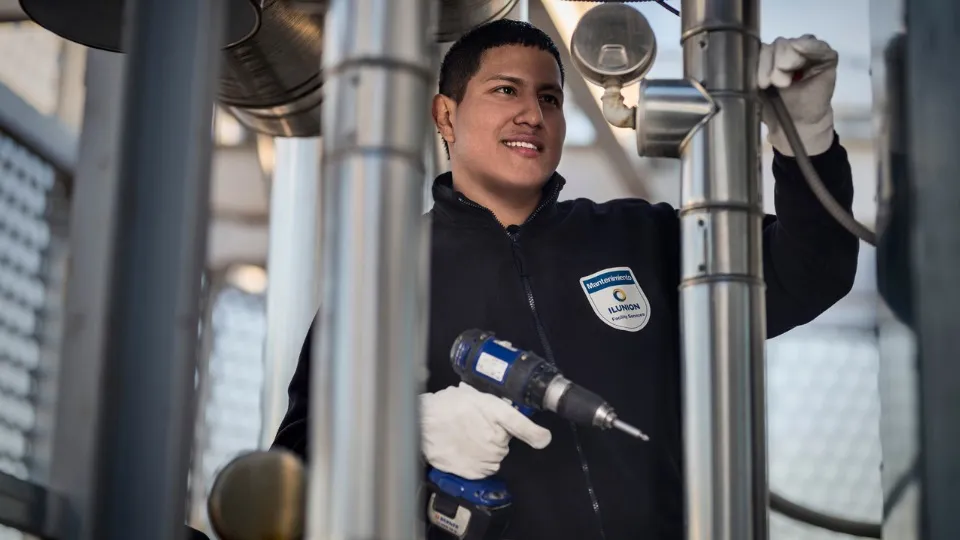 Una persona con uniforme y logotipo de ILUNION usa un taladro, sonriendo, mientras trabaja en estructuras metálicas en un entorno industrial. 
