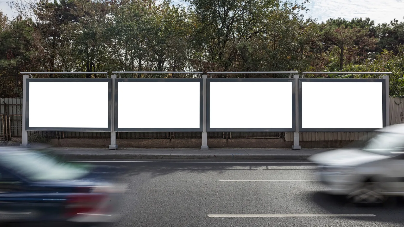 Cuatro vallas publicitarias vacías en una carretera, con sus marcos metálicos sobre un fondo de árboles frondosos y al frente una carretera con coches pasando.