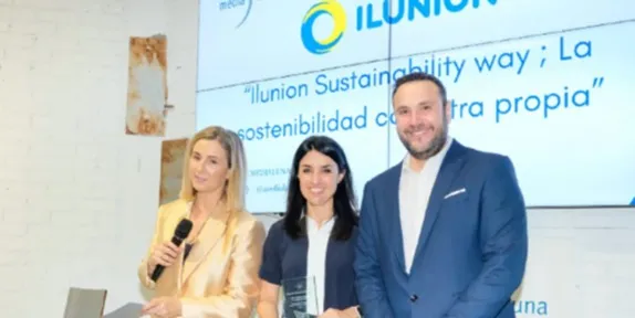 Tres personas están de pie frente a una pantalla que muestra el texto "Ilunion Sustainability way; La sostenibilidad con letra propia" durante un evento o presentación.