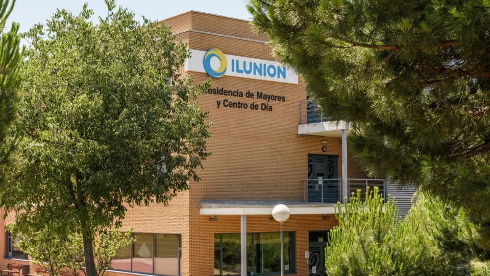 Edificio de ladrillo rojo con el cartel "ILUNION Residencia de Mayores y Centro de Día", rodeado de árboles verdes bajo un cielo azul claro, que transmite tranquilidad.