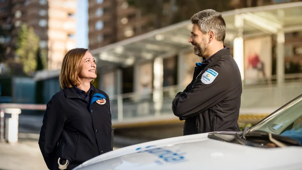 Una mujer y un hombre sonrientes, con uniformes de seguridad de ILUNION, conversan junto a un coche patrulla. El ambiente urbano sugiere un ambiente relajado y profesional.