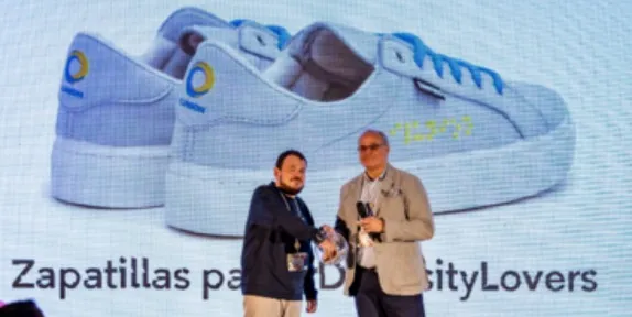 Dos hombres se dan la mano en el escenario frente a una gran imagen de zapatillas y el texto "Zapatillas para DiversityLovers" que se muestra en una pantalla.