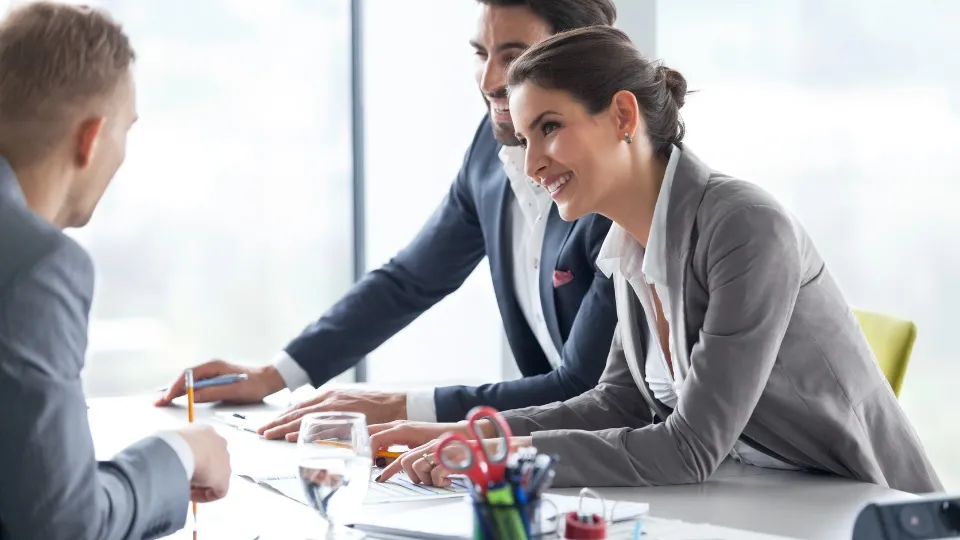Tres personas vestidas de negocios se encuentra en una reunión, sonriendo e inclinados sobre una mesa con documentos y material de oficina.