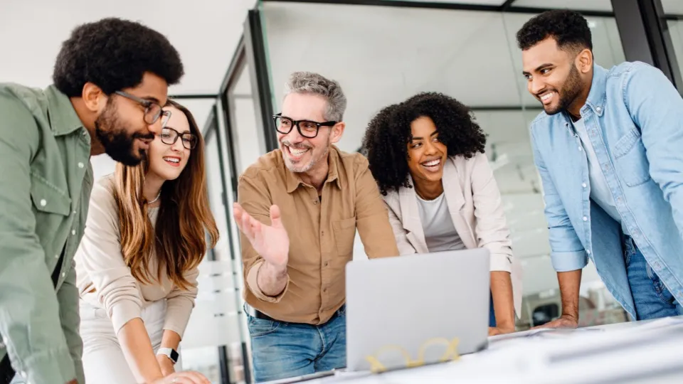 Un grupo diverso de cinco personas se reúne alrededor de una computadora portátil, sonrientes y participando en una discusión, transmitiendo un ambiente de trabajo colaborativo y positivo.