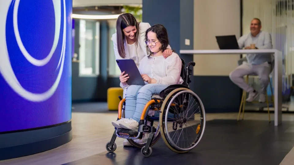 Una mujer sonriente se inclina sobre una chica en silla de ruedas, ambas mirando una tablet. Un hombre al fondo observa, sentado a una mesa con un portátil.