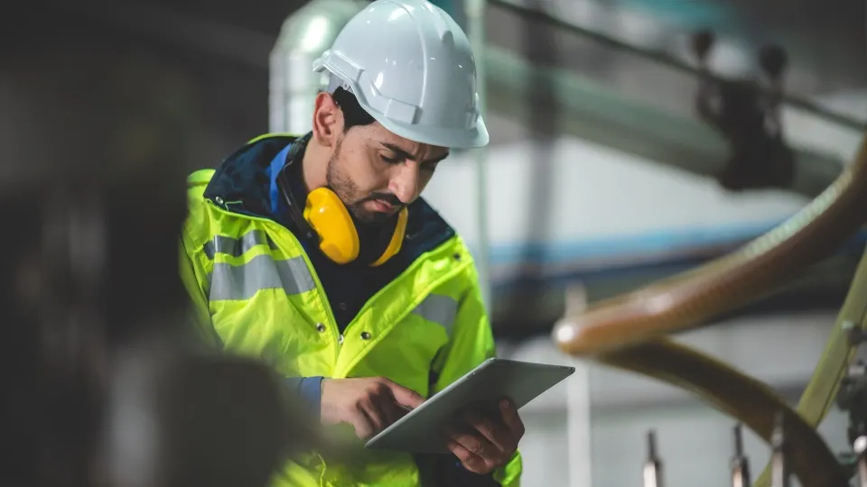 Un trabajador industrial con chaleco reflectante y casco mira fijamente una tableta. Lleva protectores auditivos alrededor del cuello en una fábrica.