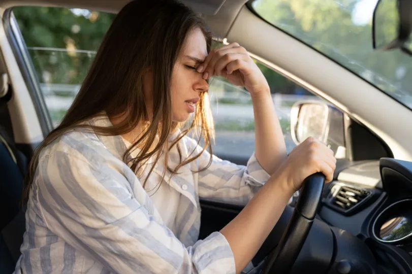 Una mujer está sentada en el asiento del conductor de un coche, sosteniendo el volante con una mano y pellizcando el puente de su nariz con la otra, pareciendo estresada o fatigada.