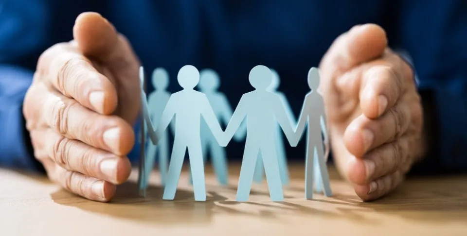 Un par de manos se ciernen protectoramente sobre un círculo de figuras de madera sobre una mesa, simbolizando protección y unidad en un contexto empresarial o comunitario.