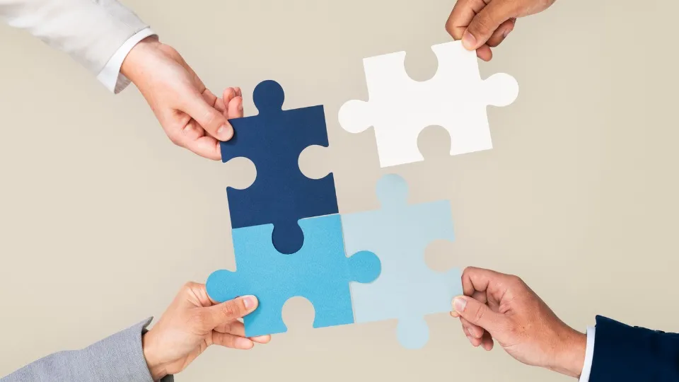 Cuatro manos sostienen piezas de rompecabezas entrelazadas en azul marino, blanco y celeste, que simbolizan el trabajo en equipo y la colaboración sobre un fondo neutro.