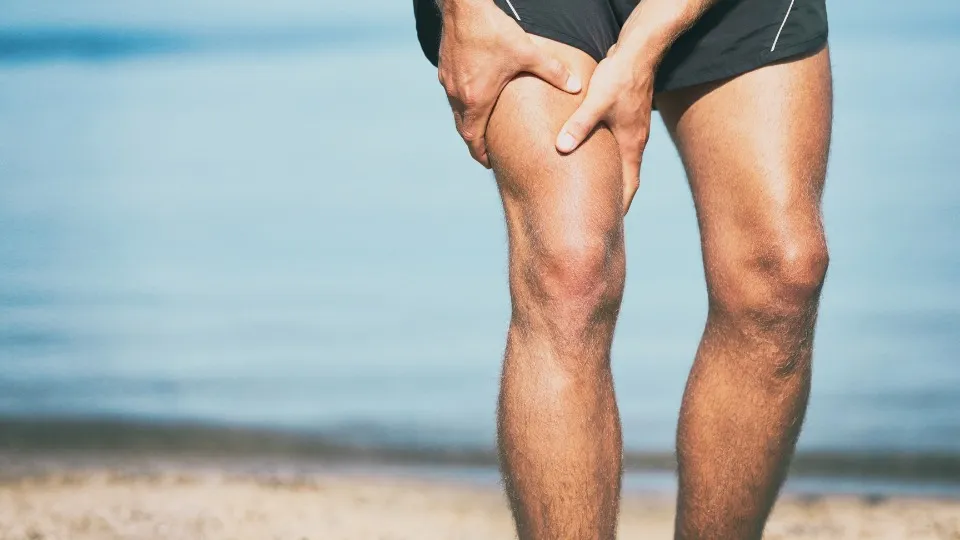 Se muestran las piernas de un hombre en la playa que se está sujetando el muslo con dolor porque se acaba de lesionar