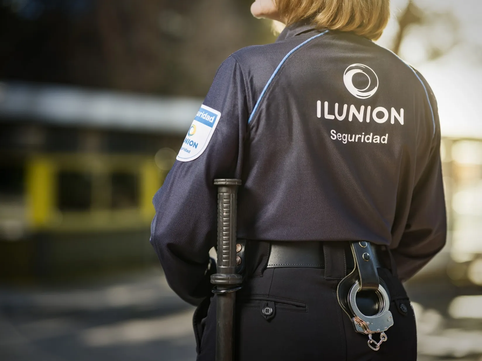 Una vigilante de seguridad de espaldas. En su uniforme se ve y se lee el logotipo ILUNION Seguridad