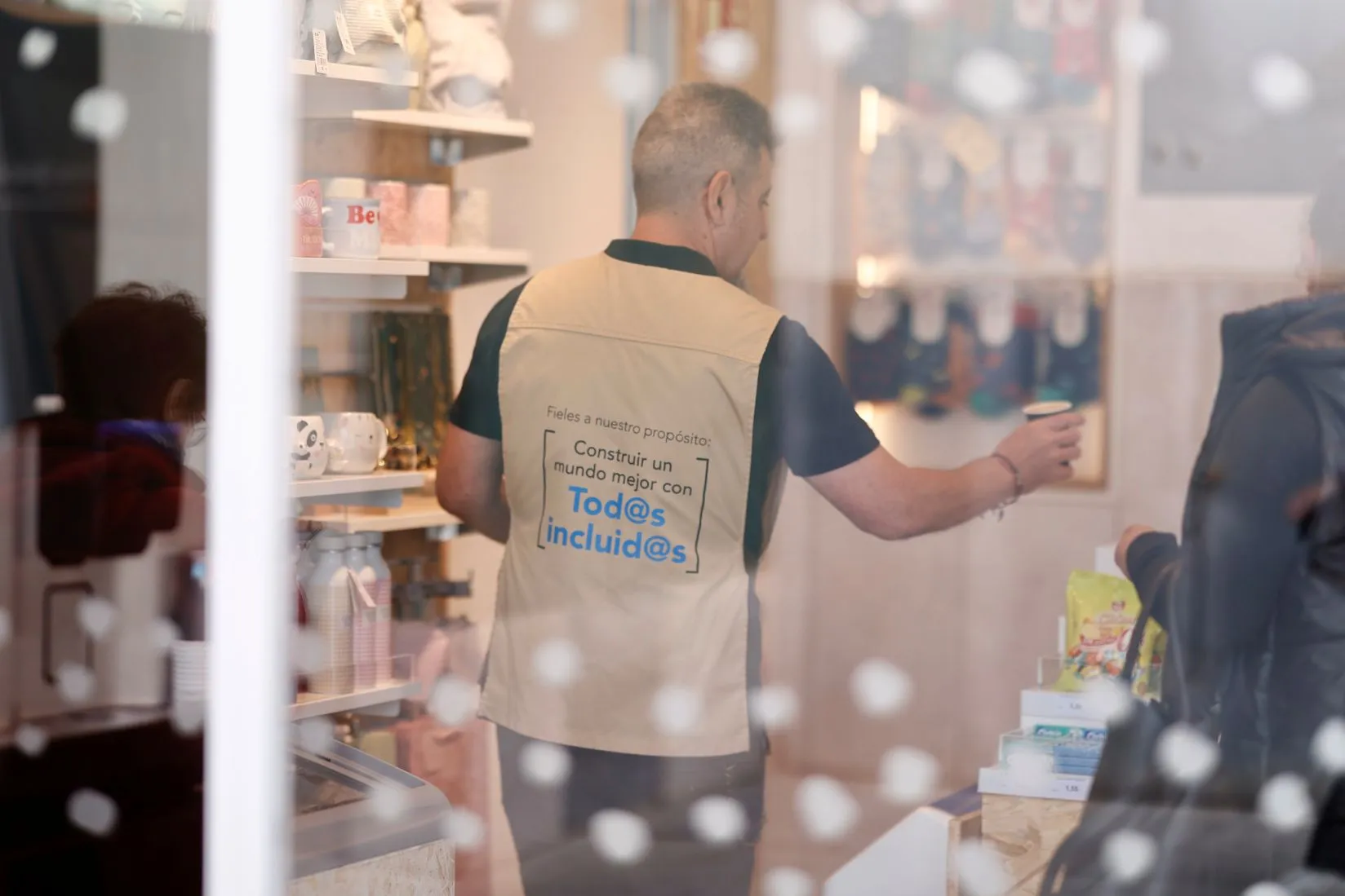 Fotografía de uno de los dependientes de la tienda, de espaldas, en su chaleco se lee todos incluidos, dando un café a una persona.