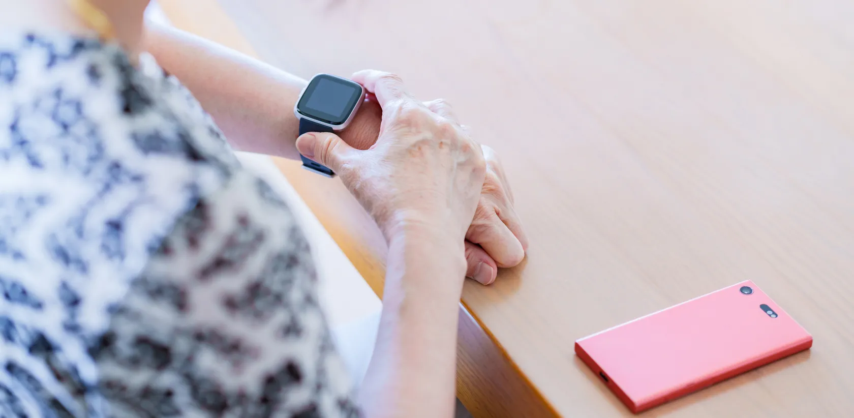 Persona con prenda de patrón blanco y negro, sentada en una mesa de madera clara, mirando un reloj inteligente en la muñeca izquierda; a la derecha hay un teléfono móvil rosa.