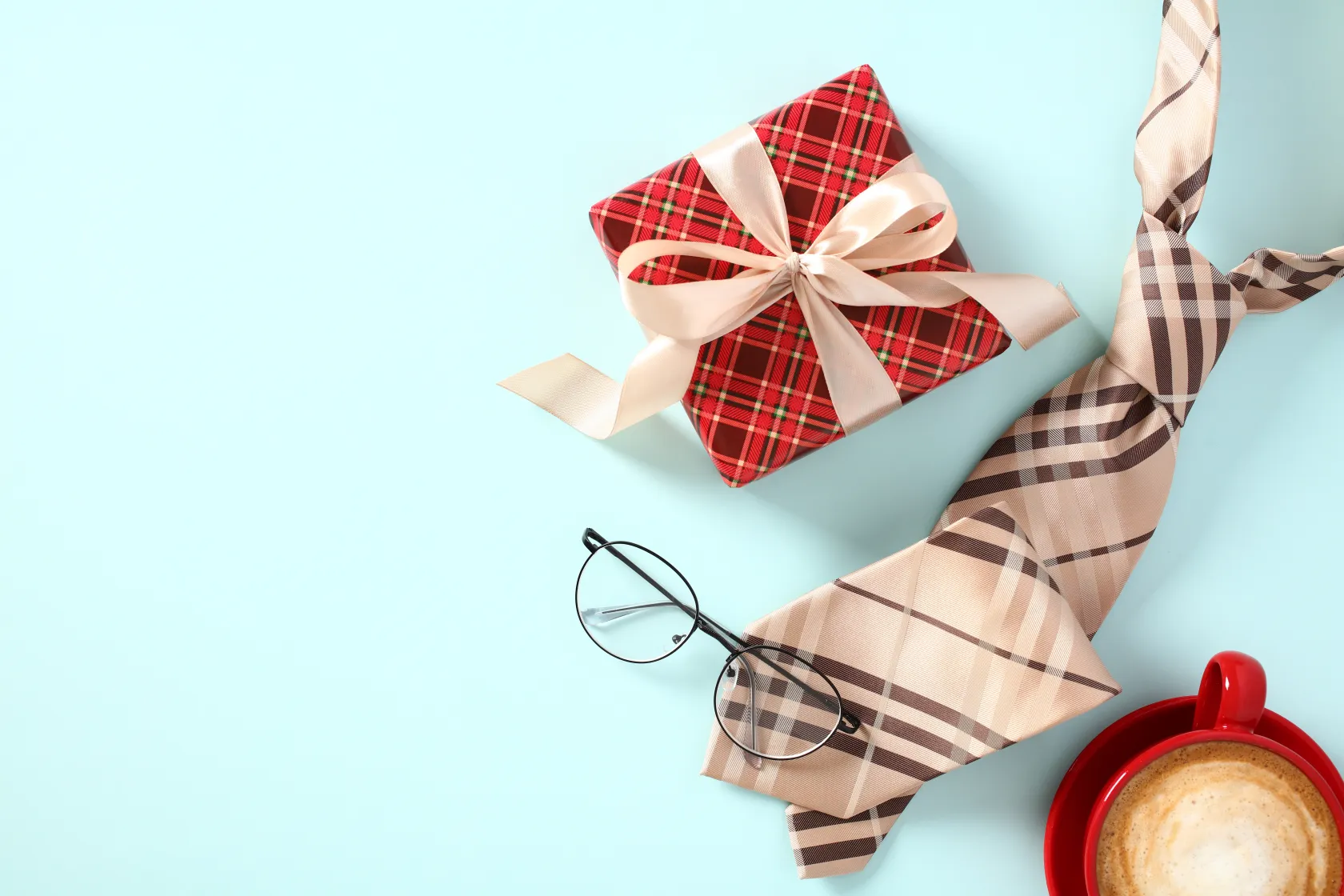 Regalo con envoltorio de estampado rojo, una corbata beige a cuadros, unas gafas y una taza de café sobre fondo celeste.
