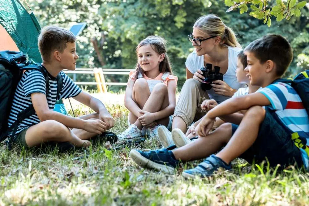 Grupo de personas sentadas en círculo sobre el césped durante una actividad al aire libre, con mochilas y una tienda de campaña al fondo