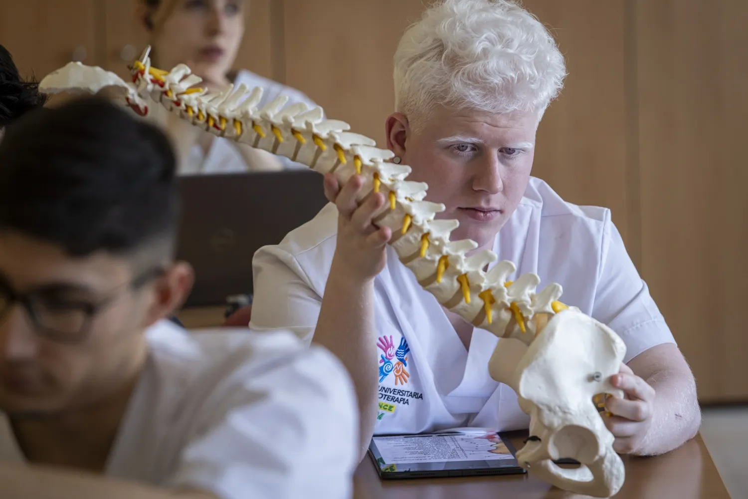 Estudiante de fisioterapia sostiene y observa un modelo de columna vertebral durante una clase práctica, con otros alumnos al fondo.