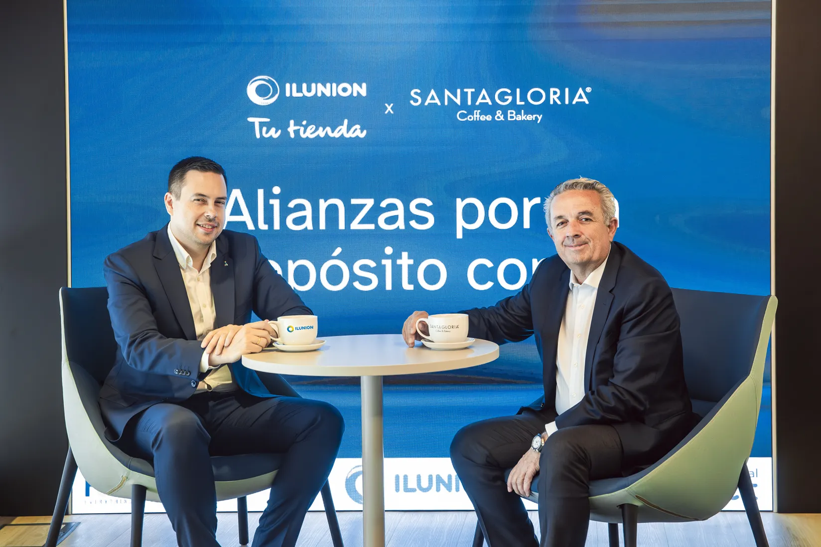 De izquierda a derecha, La firma del acuerDe izquierda a derecha, delante de un fondo azul en el que se lee 'alianzas por un propósito común', el director general de ILUNION Retail, Delio Díaz, y el presidente de FoodBox, Enrique Francia, sentados frente a una mesita con dos tazas con los logos de ILUNION y de Santagloria.