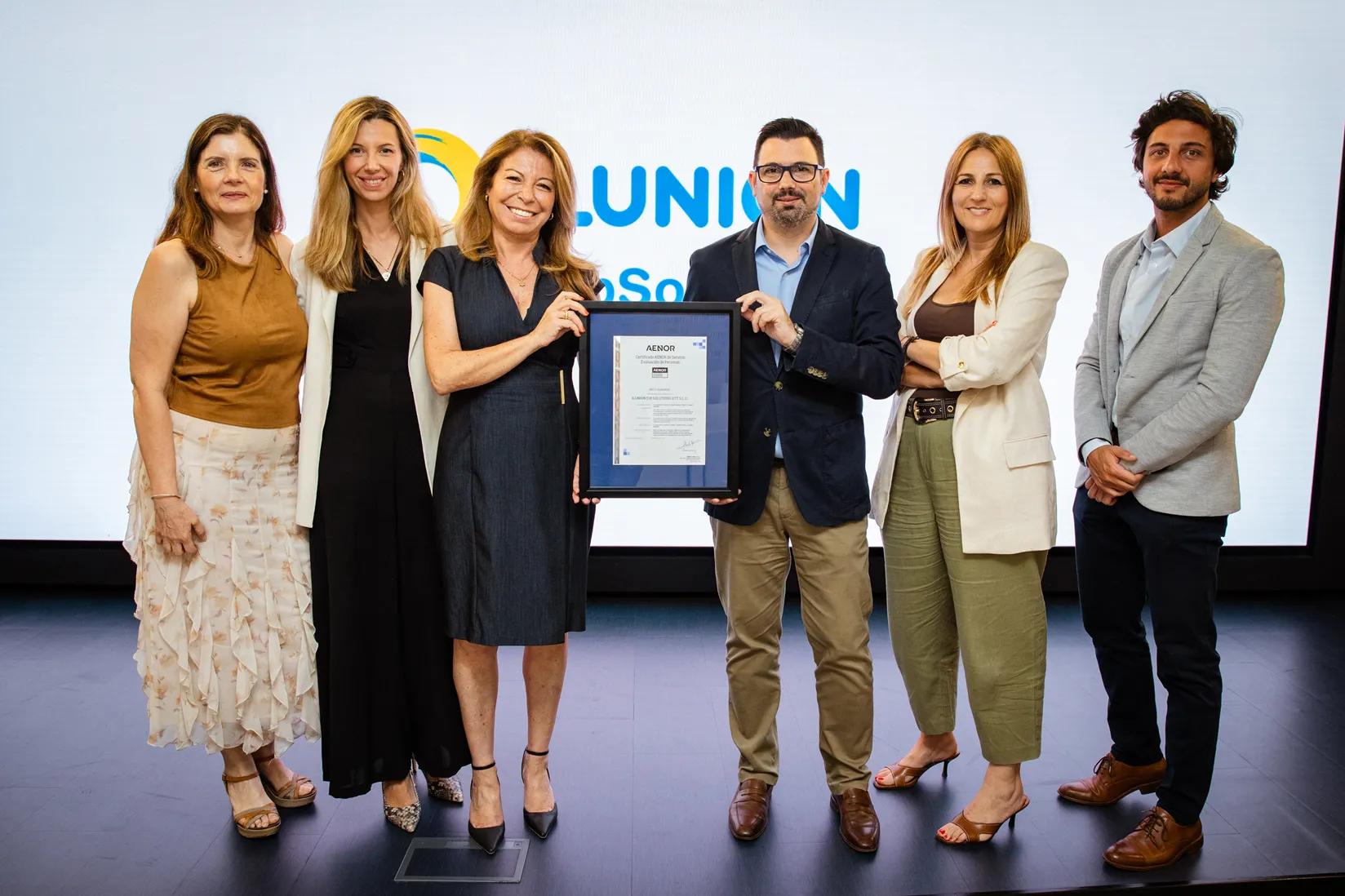 Equipo de seis personas en escenario sosteniendo un certificado AENOR frente al logotipo de ILUNION.
