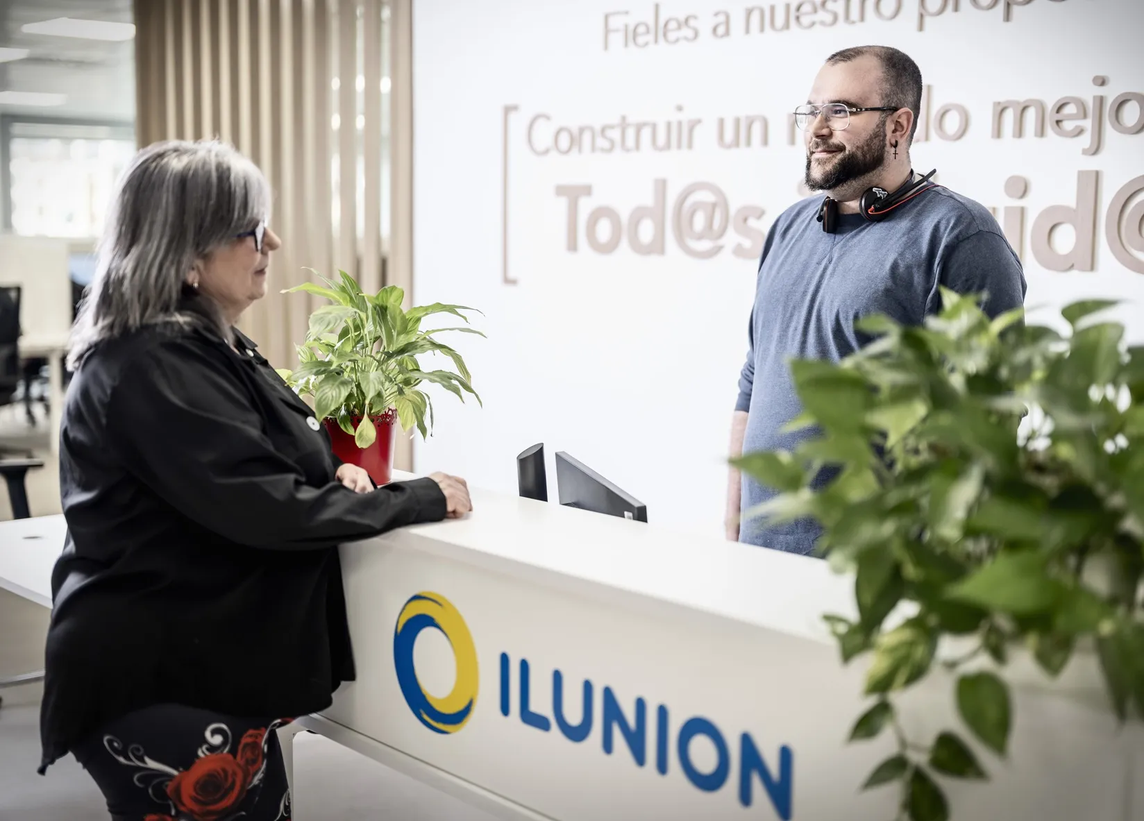 Un hombre atiende a una mujer con una muleta en un mostrador de ILUNION, en una oficina moderna con plantas decorativas y elementos de identidad corporativa visibles.
