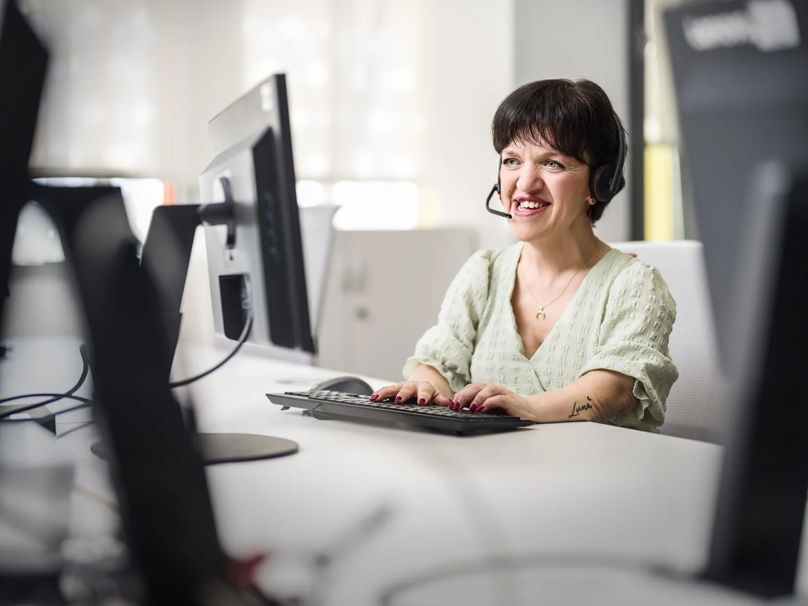 Fotografía de una mujer con auriculares y micrófono trabaja frente a un ordenador en una oficina luminosa, con las manos sobre el teclado y varios monitores alrededor.