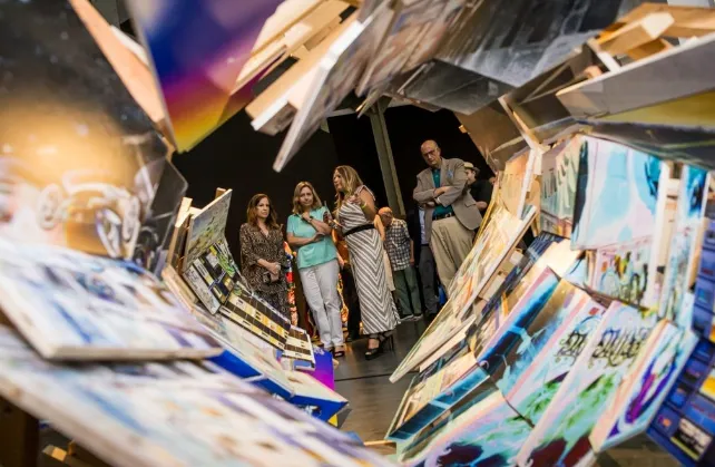 Instalación artística compuesta por numerosas obras y paneles de colores dispuestos de forma inclinada, creando un pasillo o túnel visual. Varias personas recorren y observan la exposición desde el centro del espacio. La fotografía está tomada desde el interior de la instalación, destacando la profundidad y la disposición envolvente de las piezas artísticas.
