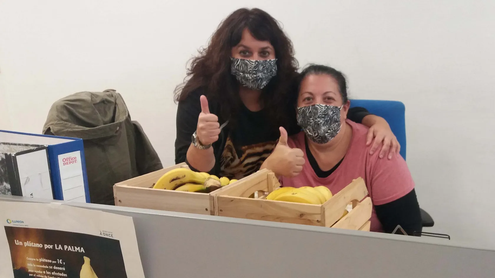 Dos trabajadoras de uno de los contact center de ILUNION posan sonrientes con el puño cerrado y el pulgar hacia arriba junto a varios plátanos